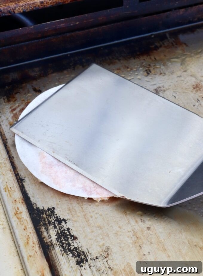 a spatula pressing a chicken burger patty, covered with parchment paper, onto a hot griddle