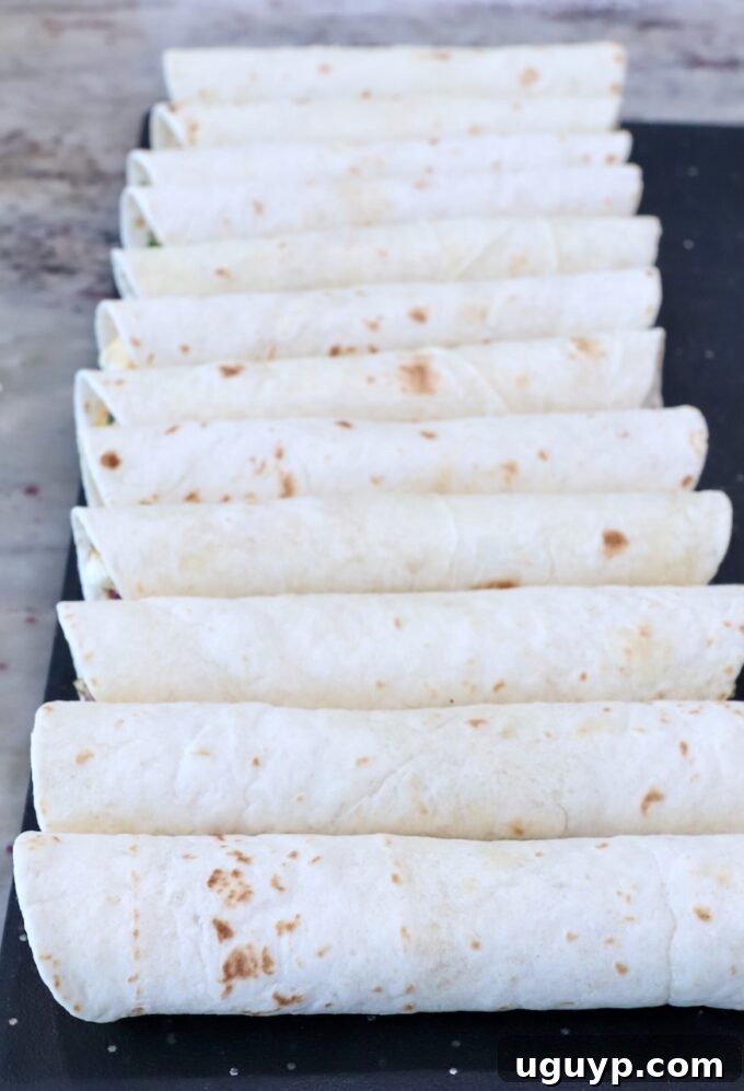 A neat row of uncooked, tightly rolled taquitos arranged on a cutting board, prepared for cooking