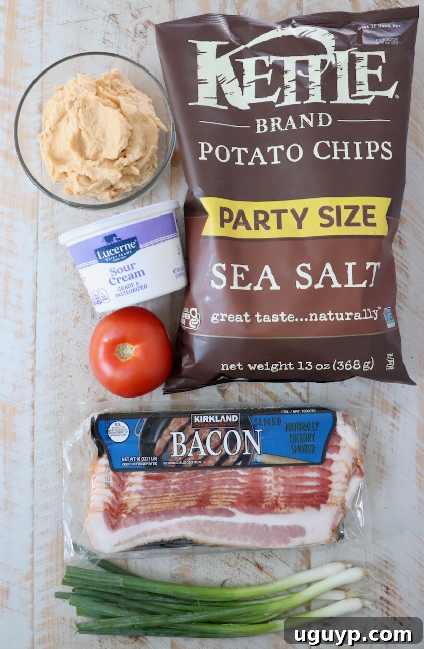 A rustic white wooden board displays the fresh ingredients laid out for making Irish nachos, including golden potato chips, vibrant green onions, diced red tomatoes, creamy sour cream, and crispy bacon bits.