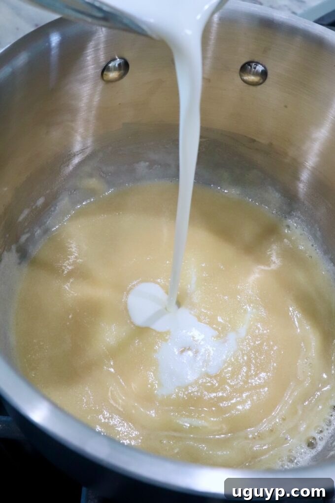 Heavy cream being slowly poured into a golden roux in a saucepan on the stove, forming the base of a creamy sauce.