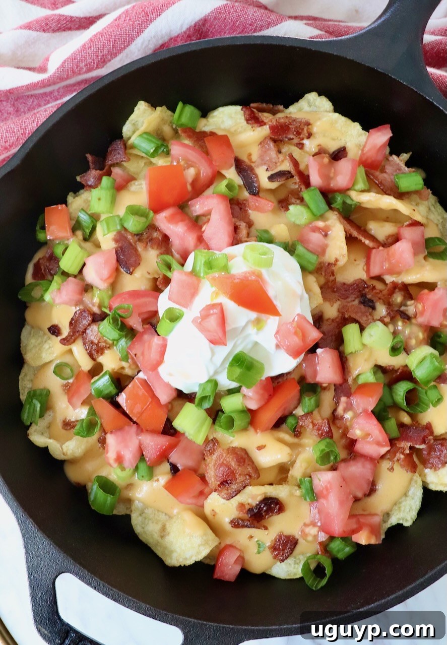 A vibrant cast iron skillet of Irish nachos, topped generously with creamy beer cheese sauce, crispy bacon, fresh green onions, and diced tomatoes, with a perfect dollop of sour cream in the center.