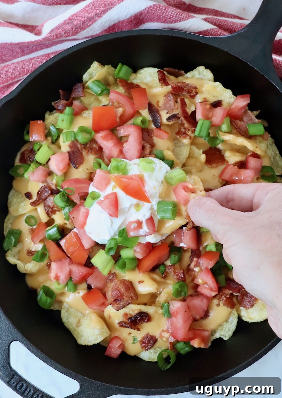 A hand reaching into a cast iron skillet of Irish nachos to dip a chip into the creamy sour cream, highlighting the delicious textures and inviting nature of the dish.