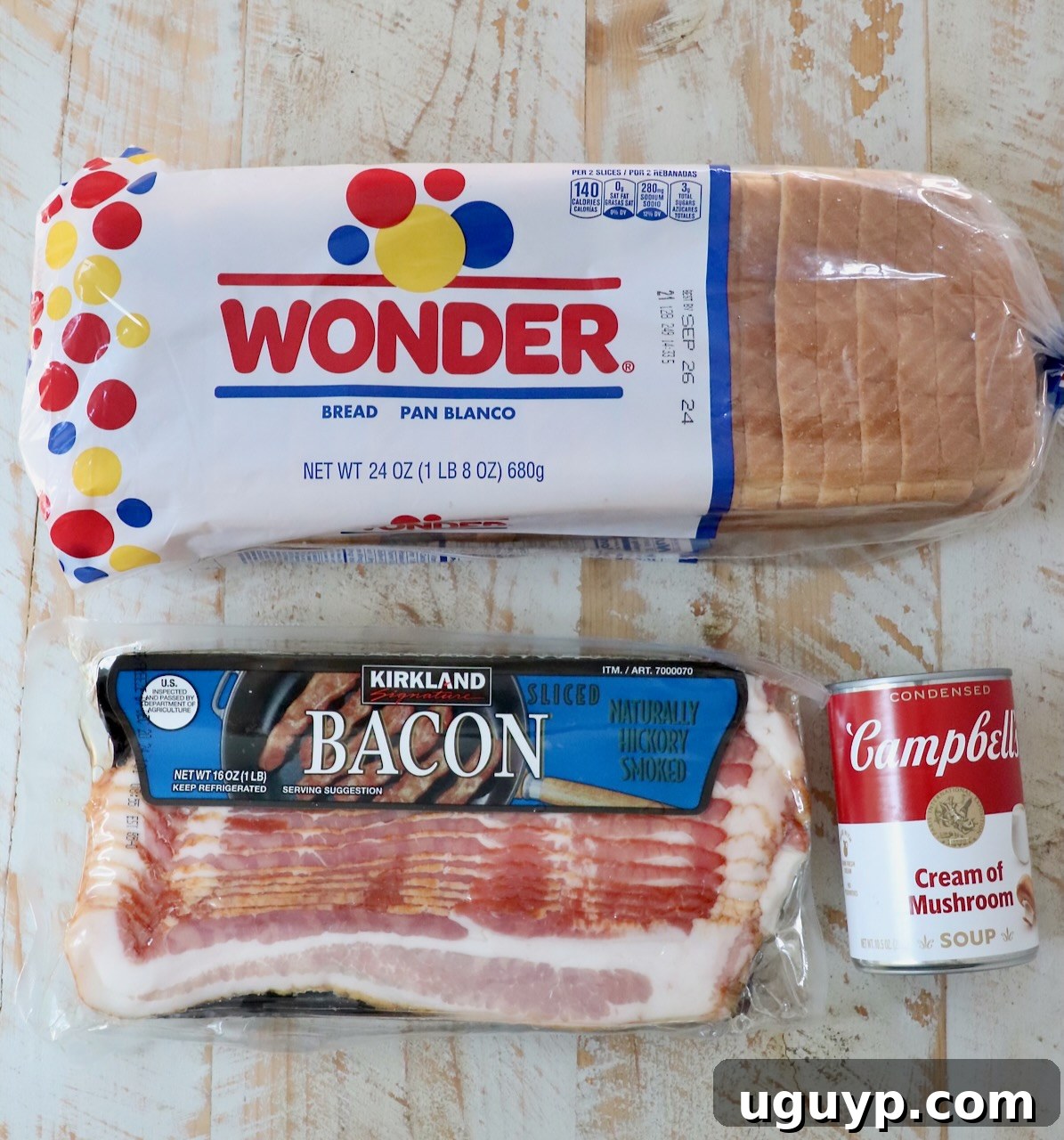 Ingredients for Bacon Roll Ups: a package of regular sliced bacon, a loaf of white sandwich bread, and a can of condensed cream of mushroom soup displayed on a rustic white wooden board.