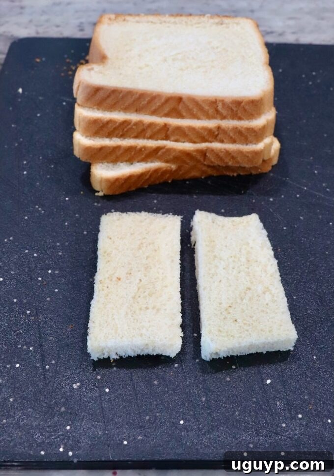 Stacked white bread slices on a cutting board, with one piece cut in half and crusts removed, demonstrating the first step of preparation.