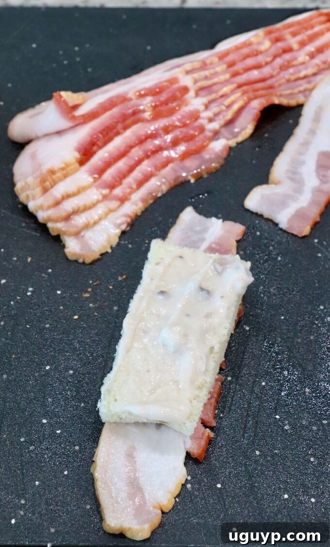 A slice of white bread evenly coated with cream of mushroom soup, placed on a half strip of bacon, ready for rolling.