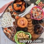 A festive Halloween snack board with small bowls of pretzels, popcorn, candies, and cookies, all arranged on a round wooden board for a party.
