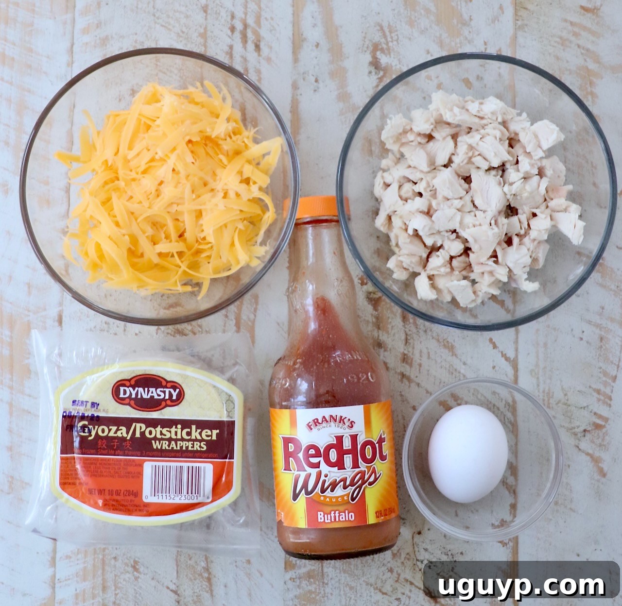 A flat lay of the essential ingredients for Buffalo Chicken Bites laid out on a white wooden board, including shredded chicken, buffalo sauce, cheese, egg, and potsticker wrappers.