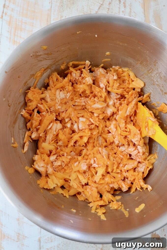 Shredded cooked chicken being tossed with buffalo sauce in a mixing bowl using a spatula, creating a vibrant orange mixture.