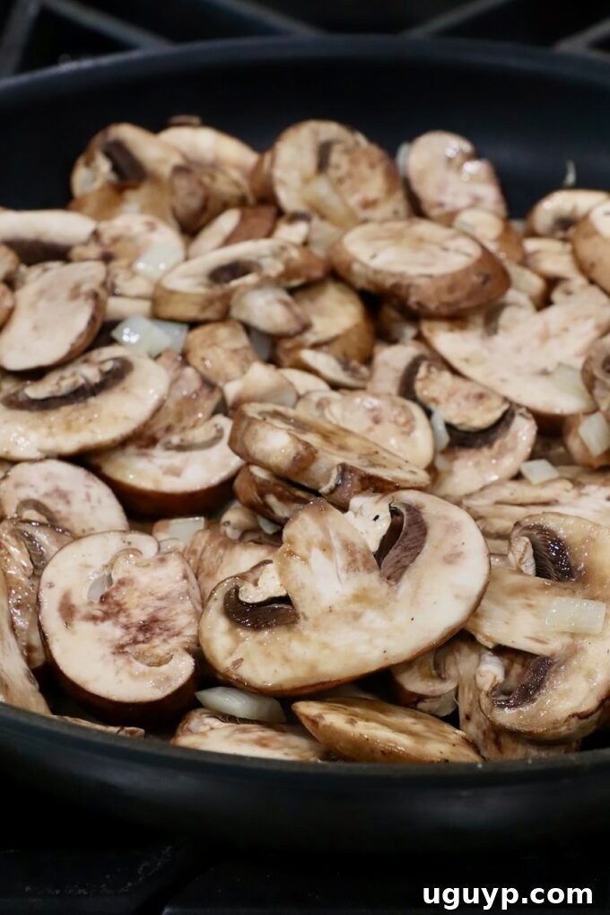 Sliced baby bella mushrooms beginning to cook in a large skillet with olive oil.