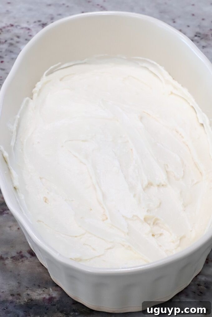 Smooth and creamy cream cheese filling spread evenly in the bottom of a casserole dish, forming the first layer of the dip.