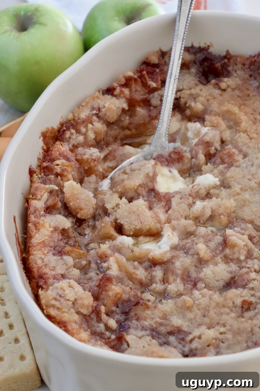 A close-up of the warm, baked apple pie dip in a casserole dish, with a serving spoon scooping out a generous portion, highlighting its delicious layers.