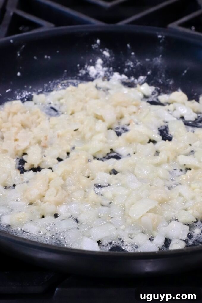 A large skillet containing sautéed diced onions and minced garlic, with all-purpose flour sprinkled evenly over them, ready to form a roux.
