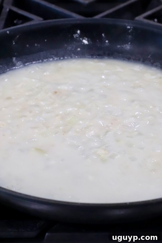 A creamy sauce in a skillet, thickened with flour and chicken broth, featuring visible diced onions and garlic, simmering gently.