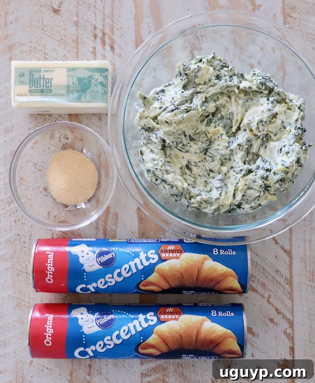 Ingredients for spinach pinwheels laid out neatly on a white wood board, including crescent roll dough, spinach artichoke dip, butter, and garlic powder.