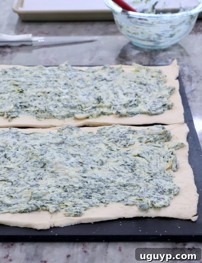 Spinach artichoke dip being spread evenly over an unrolled crescent roll dough sheet on a cutting board.