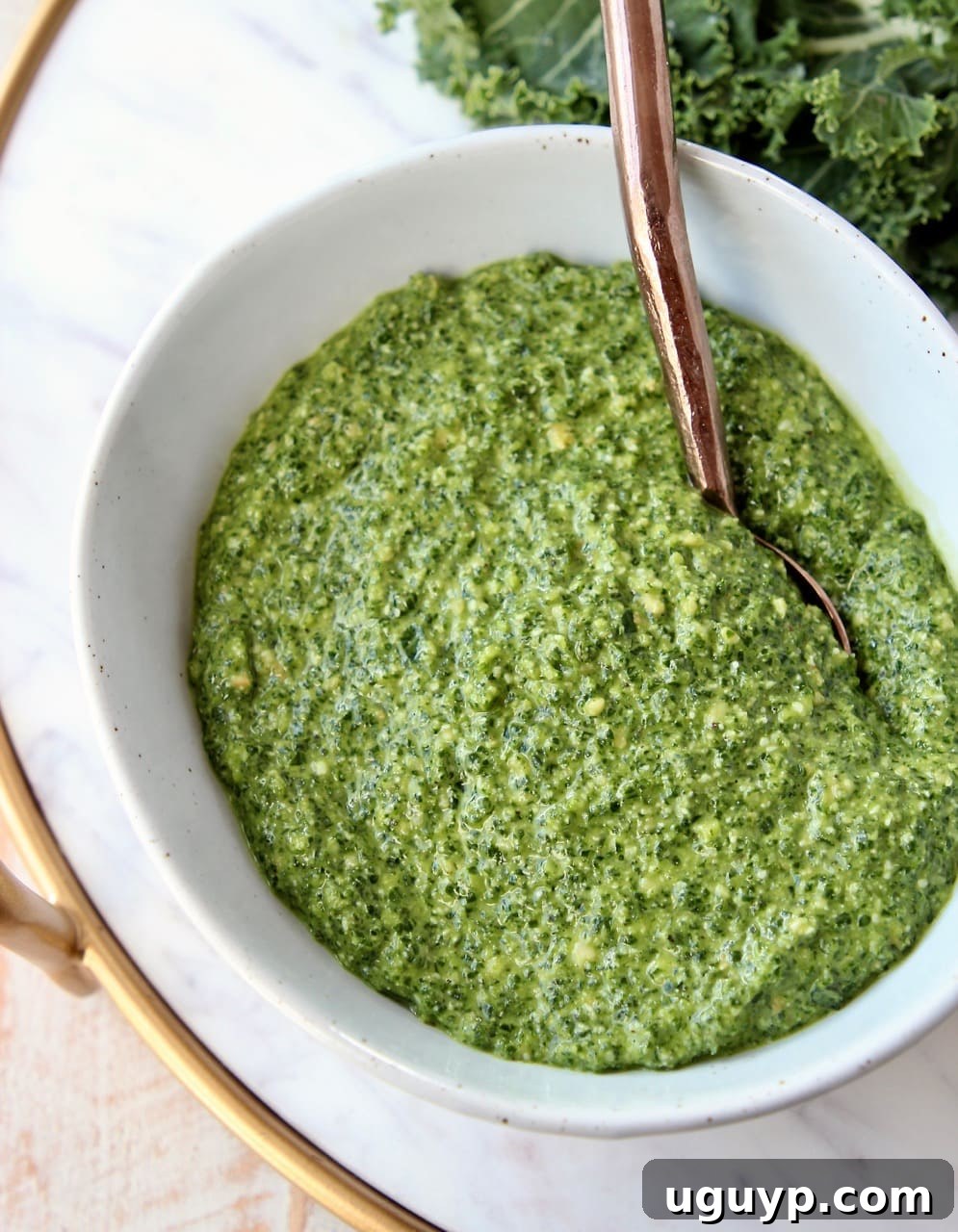 Freshly made kale pesto in a white ceramic bowl with a copper spoon resting in it, showcasing its rich green color and smooth texture.