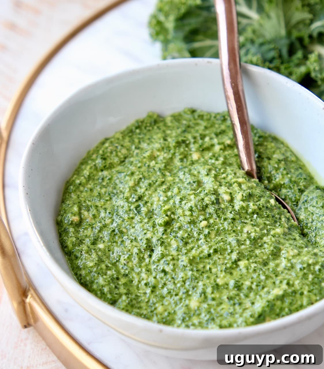 A close-up of kale pesto in a white bowl, with a copper spoon scooping out a generous portion, highlighting its texture.