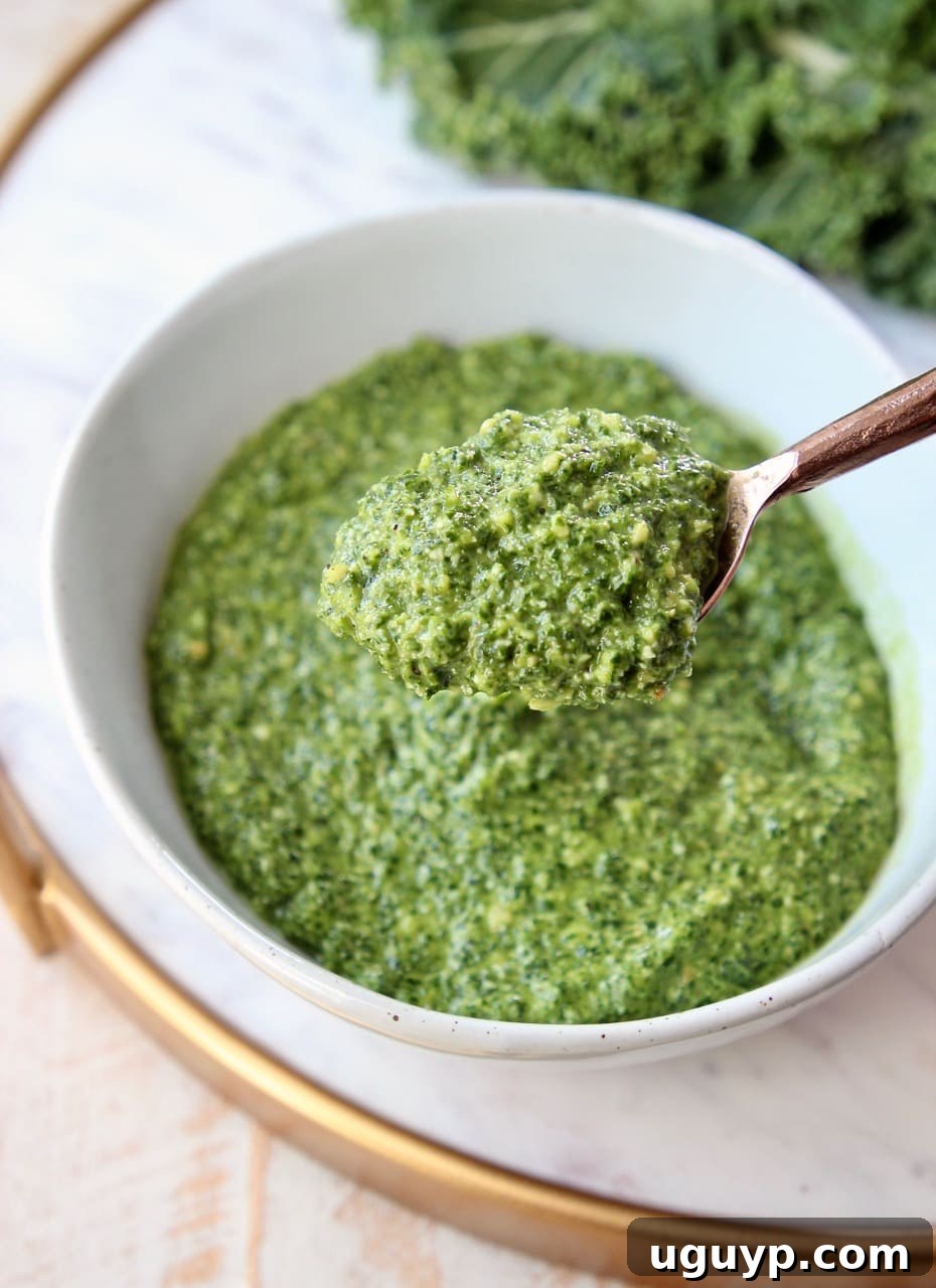 A copper spoon pulling out a dollop of vibrant green kale pesto from a white bowl, showing its thick texture.