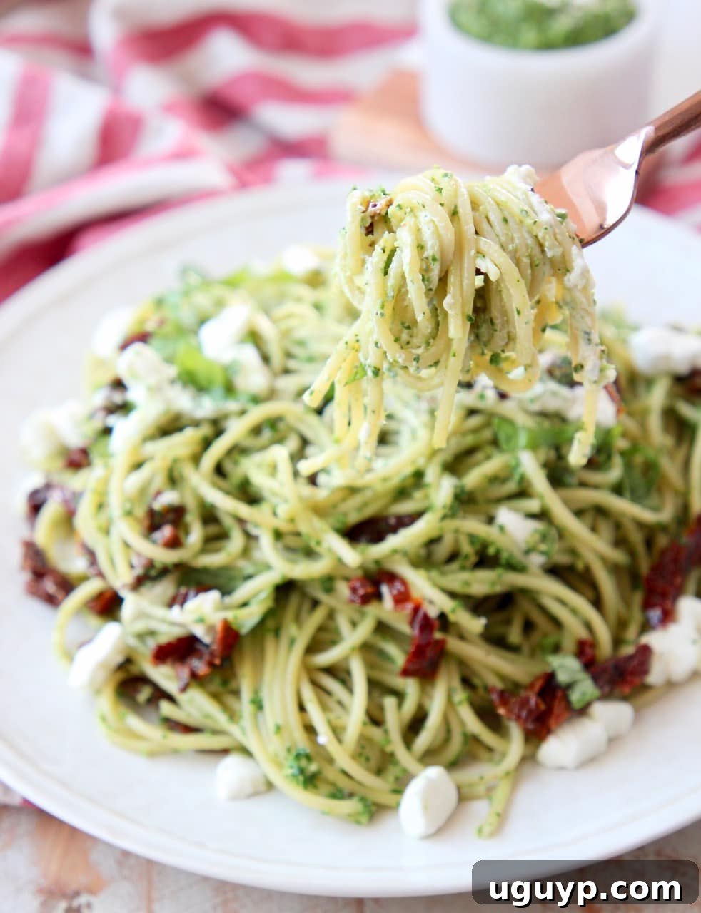 Pesto spaghetti elegantly wrapped around a copper fork, held over a plate adorned with more pesto spaghetti, crumbled goat cheese, and vibrant sun-dried tomatoes.