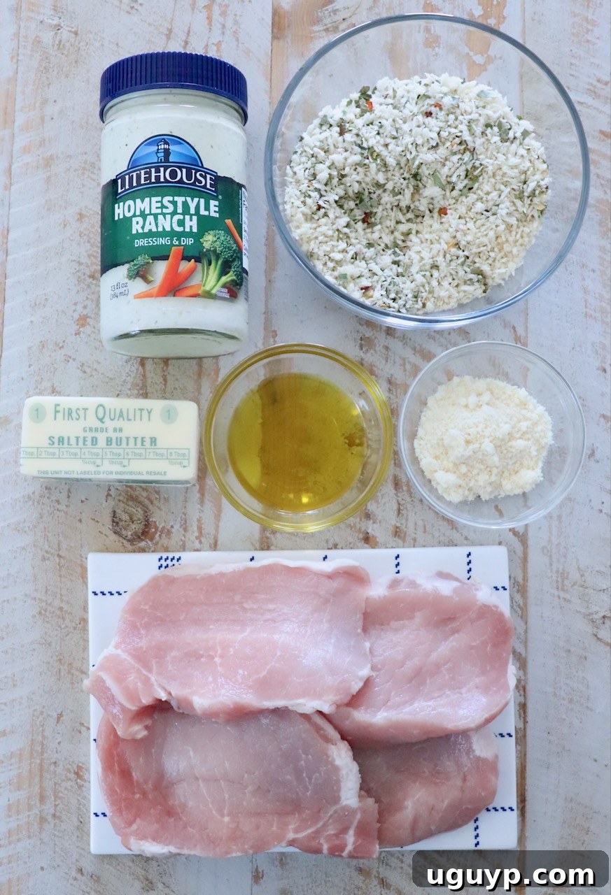 Ingredients for breaded pork chops, including boneless pork chops, ranch dressing, breadcrumbs, parmesan, butter, and olive oil, laid out on a white wooden board.