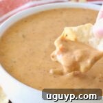 Hand dipping a tortilla chip into chili cheese dip in a bowl, ready to eat.