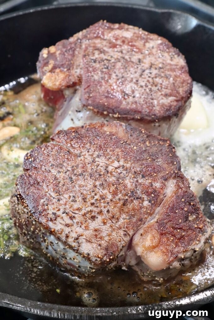 A cast iron skillet with two seared steaks, surrounded by melting butter and garlic cloves on the stove.