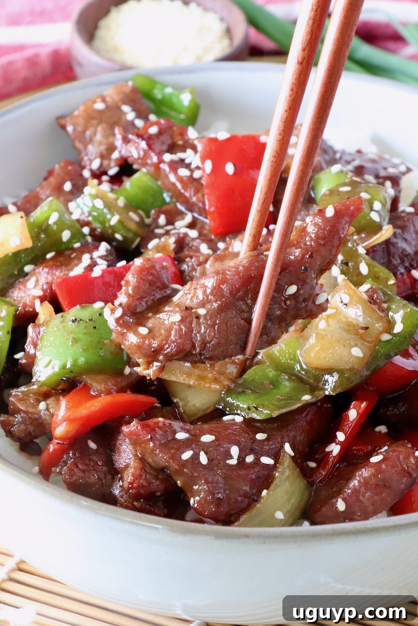 Chopsticks holding a piece of tender steak over a bowl of Chinese Pepper Steak Stir Fry with vibrant peppers and onions.