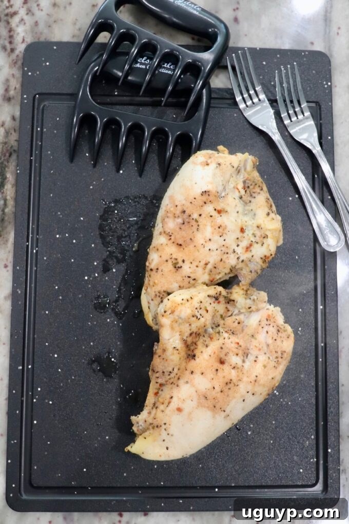 Perfectly cooked and tender chicken breasts on a cutting board, flanked by two forks and meat claws, poised for easy shredding.