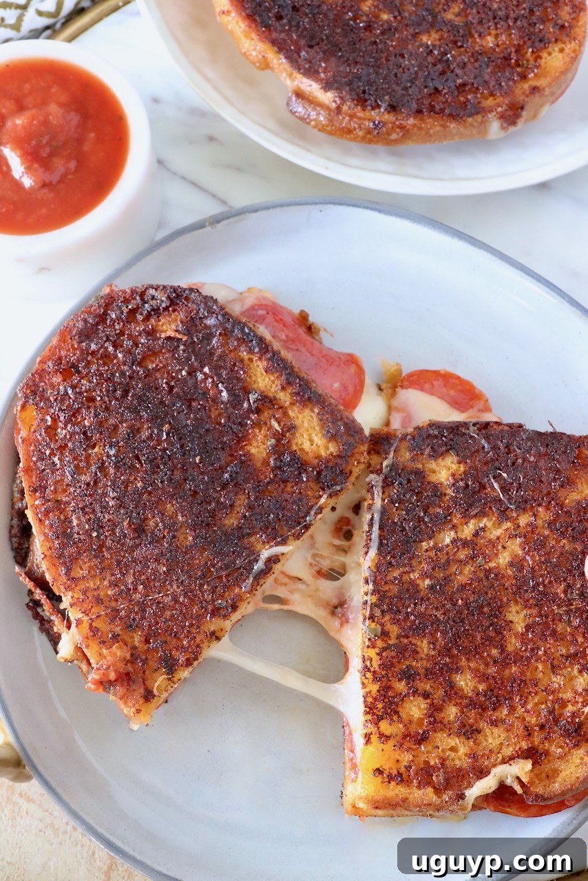 A Pizza Grilled Cheese sandwich, perfectly cut in half and pulled apart to show the melted cheese, next to a small bowl of savory pizza sauce for dipping, on a white plate.