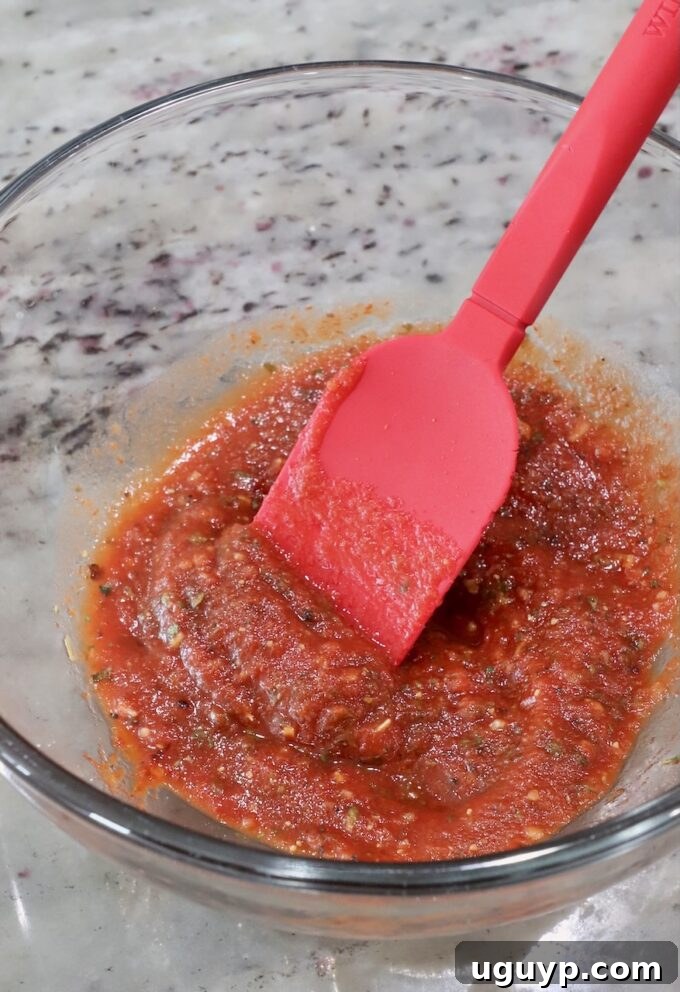 Rich red pizza sauce generously seasoned with aromatic pizza seasoning, stirred in a glass bowl with a red silicone spatula, awaiting assembly.