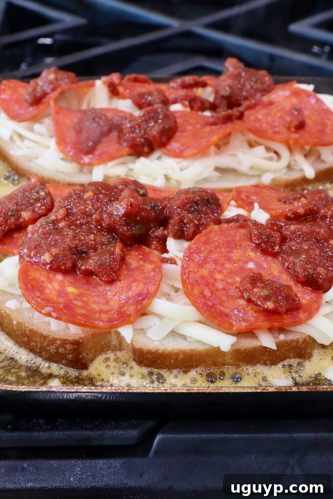 Two slices of buttered sourdough bread cooking in a hot skillet, topped with melted mozzarella cheese, savory pepperoni slices, and a dollop of seasoned pizza sauce, ready for the top bread slice.