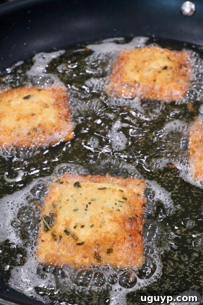 Several breadcrumb-coated raviolis sizzling to a golden crisp in a skillet filled with hot oil on a stove, showcasing the pan-frying process.