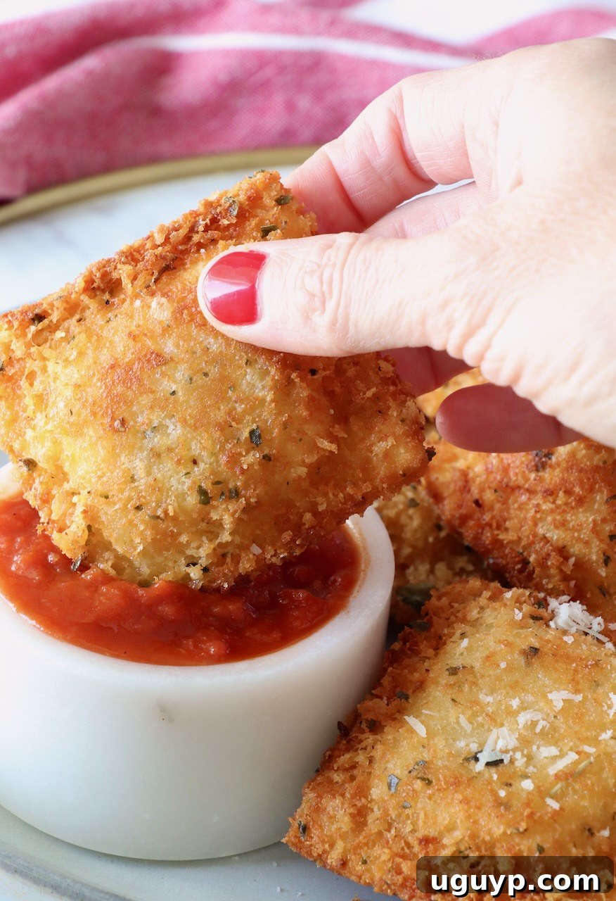 A close-up of a hand holding a perfectly toasted ravioli, gently dipping it into a small, inviting bowl of vibrant marinara sauce.
