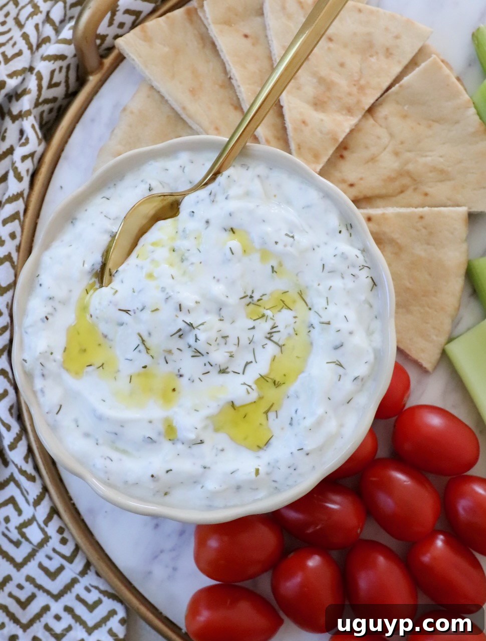 Tzatziki sauce in a white bowl with a spoon, garnished with a swirl of olive oil and a sprinkle of fresh dill. Slices of pita bread and vibrant cherry tomatoes are arranged beside the bowl.