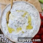 tzatziki sauce in a bowl with a spoon with olive oil drizzled on top and slices of pita bread and cherry tomatoes on the side