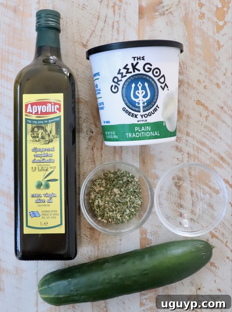 The essential ingredients for making homemade tzatziki sauce are neatly arranged on a rustic white wooden board, including a whole English cucumber, a container of Greek yogurt, a bottle of olive oil, a bottle of white vinegar, and a small bowl of Greek seasoning blend.