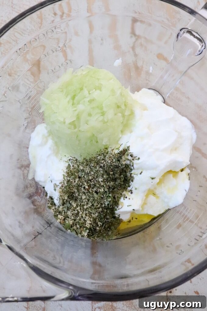 A large clear glass bowl contains plain Greek yogurt, freshly grated cucumber (partially drained), a drizzle of olive oil, and a generous amount of Greek dried herbs and spices, ready to be mixed for tzatziki sauce.