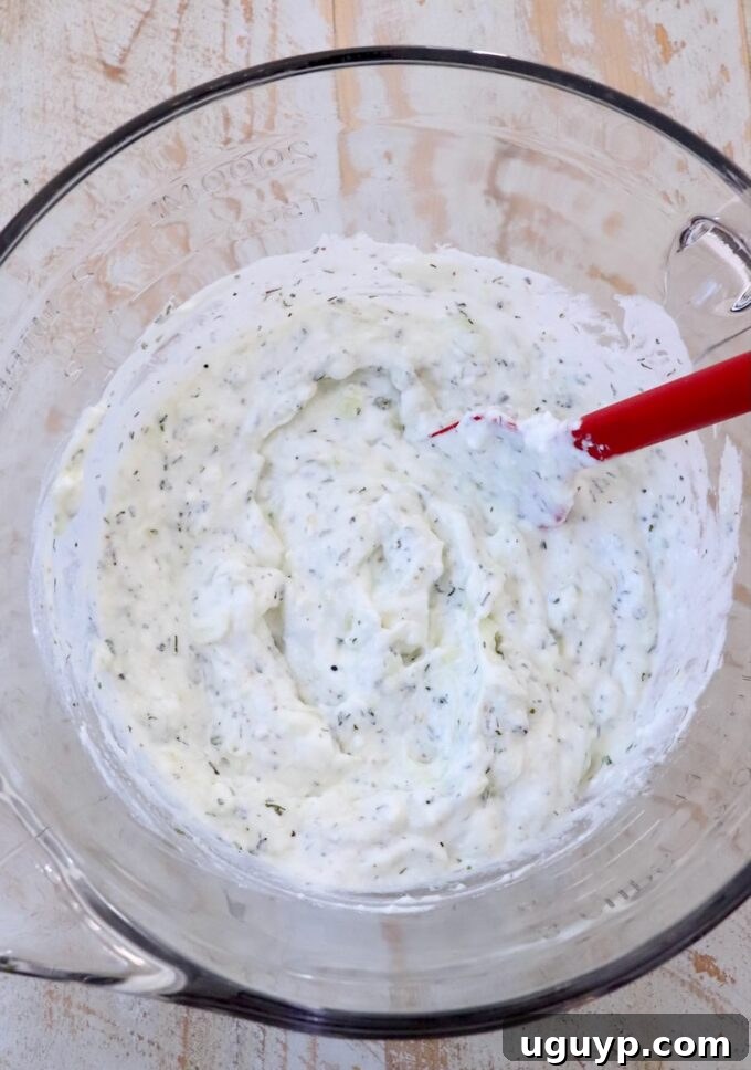 The tzatziki sauce is thoroughly mixed in a large clear glass bowl with a red spatula, showcasing its creamy, consistent texture with flecks of cucumber and herbs.