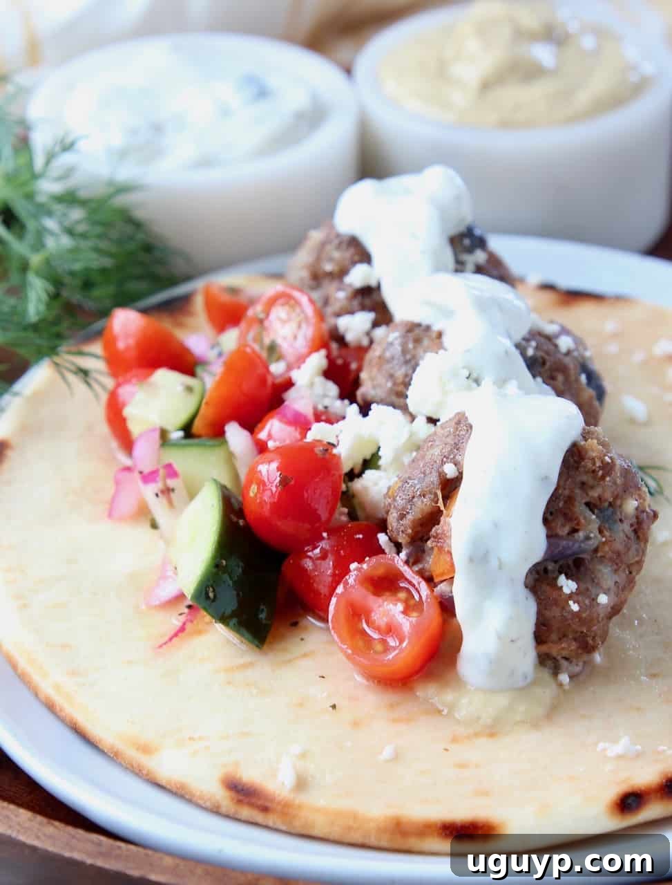 Three Greek meatballs are nestled on warm pita bread, generously topped with a fresh tomato cucumber salad and a dollop of creamy tzatziki sauce. The setting evokes a delicious and wholesome Mediterranean meal.