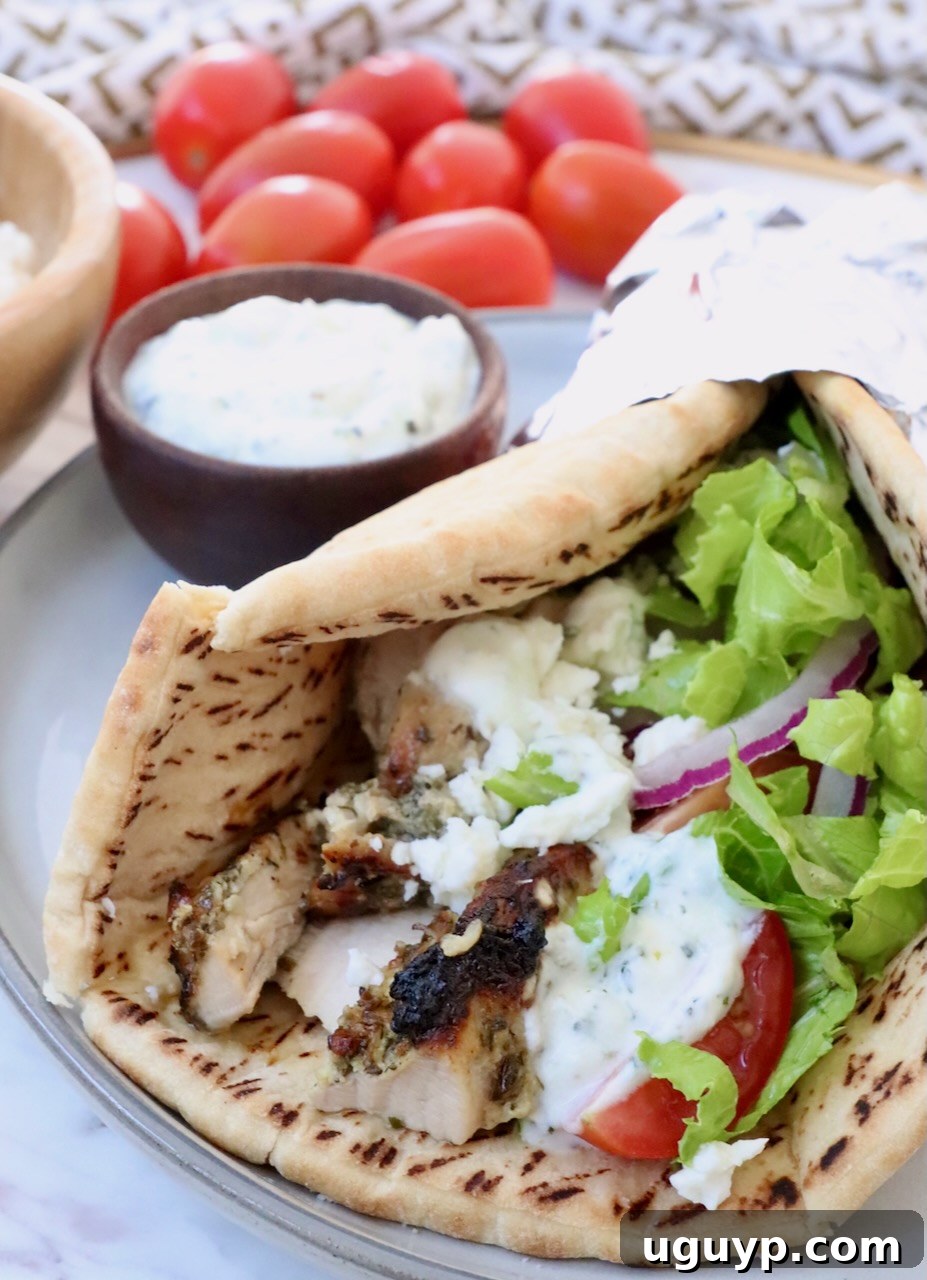 A beautifully composed Greek chicken gyro, showcasing thinly sliced marinated chicken meat, creamy tzatziki sauce, crisp shredded lettuce, and fresh slices of onion and tomato, all nestled in warm pita bread and presented on a plate.