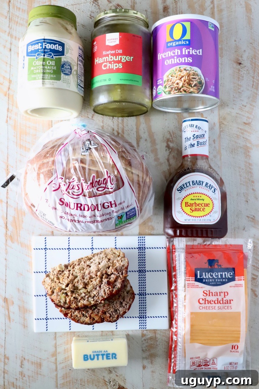 Ingredients for a meatloaf sandwich laid out on a white wooden board, including bread, meatloaf slices, cheese, pickles, and sauces.