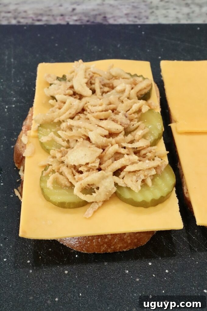 One slice of bread on a cutting board, layered with cheese, dill pickle slices, and fried onion strings.
