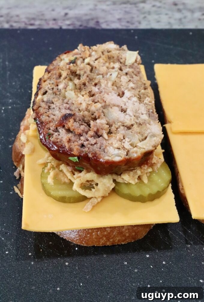A slice of cooked meatloaf placed on top of the layered cheese, pickles, and fried onion strings on a piece of bread.