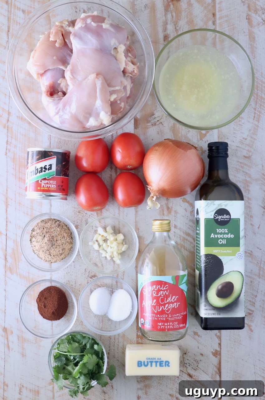 Ingredients for Mexican Chicken Adobo laid out on a white wooden board, including chicken thighs, tomatoes, onions, chipotle peppers, and spices.