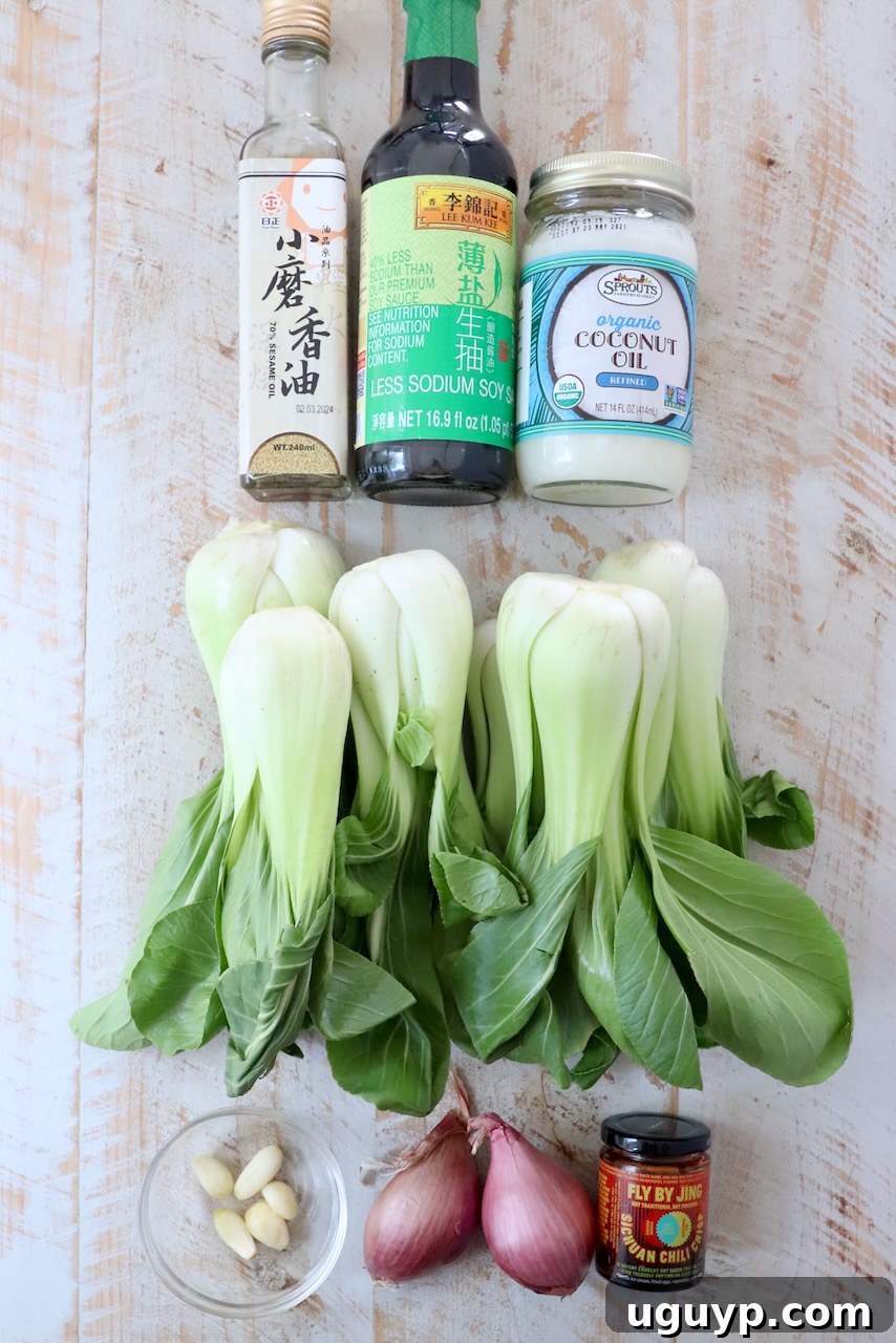 A selection of fresh ingredients for baby bok choy arranged neatly on a rustic white wood board, including whole bok choy, garlic, shallots, soy sauce, and sesame oil.