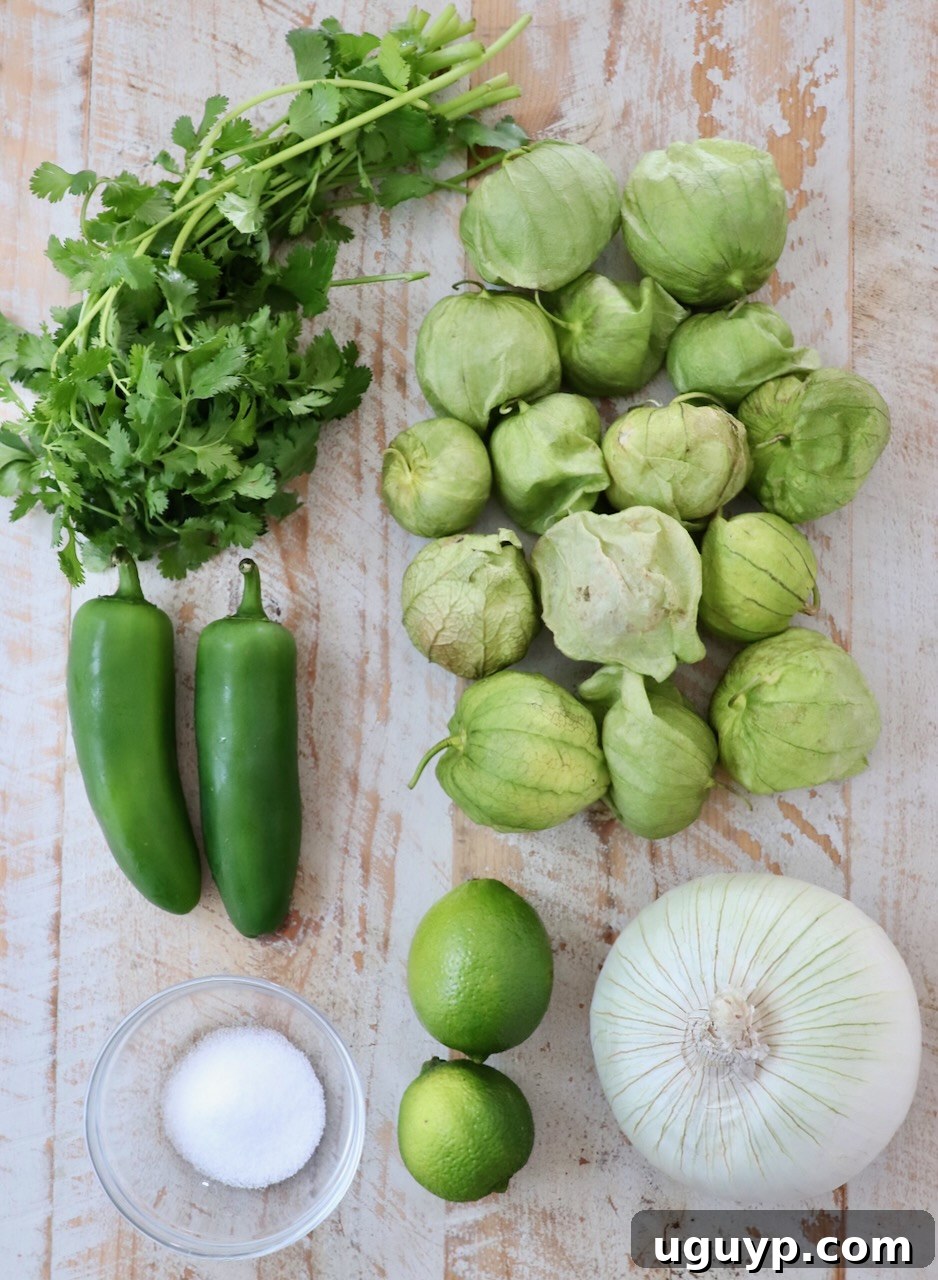 Fresh ingredients for tomatillo salsa laid out on a white wooden board, including tomatillos, jalapeños, onion, and cilantro.