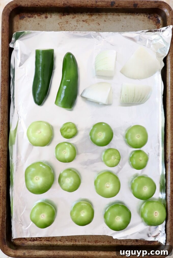Fresh tomatillos, white onion quarters, and whole jalapeños arranged on a foil-lined baking sheet before roasting.