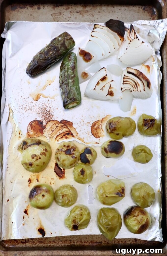 Roasted tomatillos, white onion, and jalapeños on a baking sheet, showing a slight char and softened appearance.