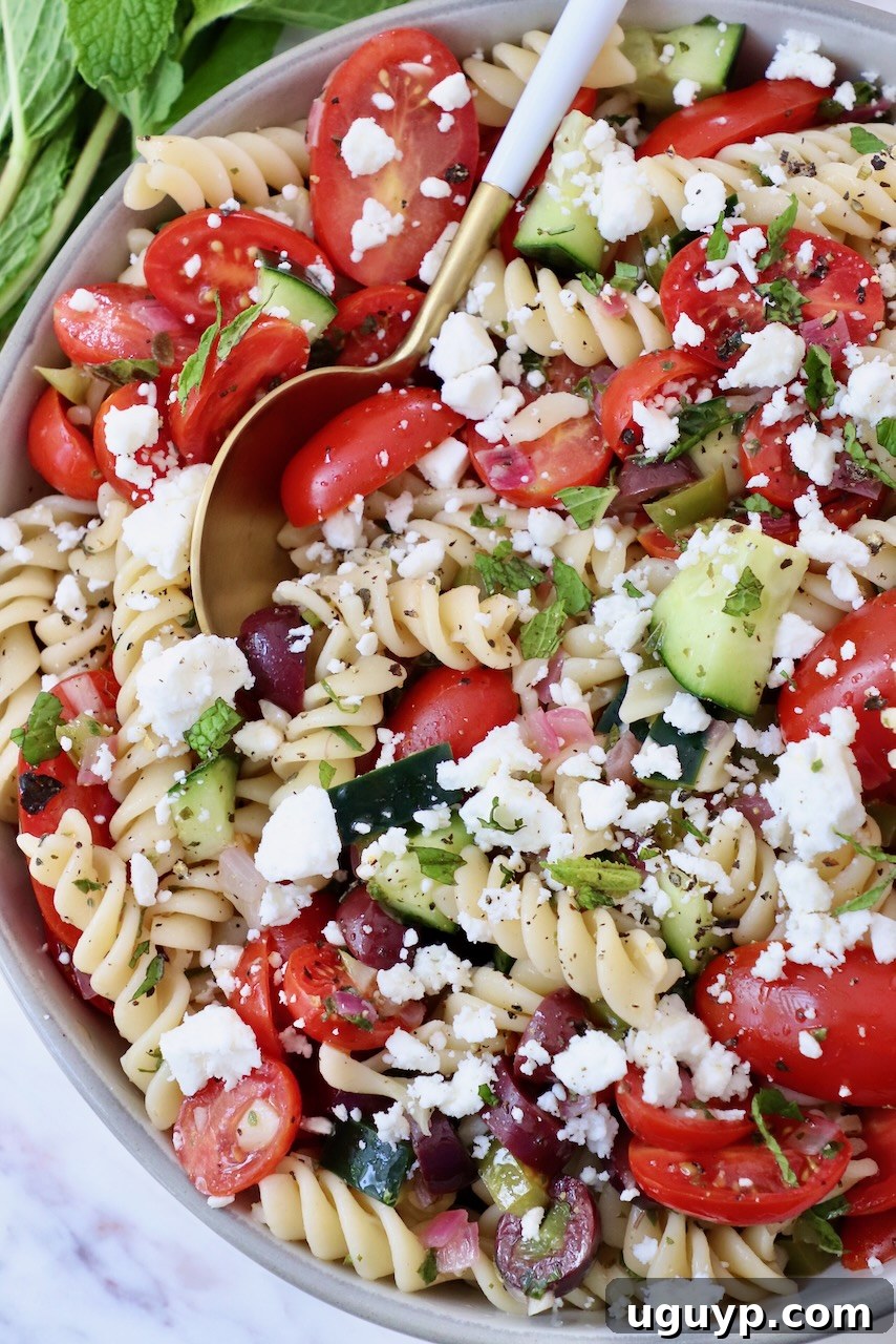 A large bowl of Greek pasta salad with cucumber, tomatoes, olives, and feta, ready to be served with a spoon.