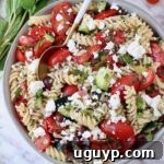 A bowl of Greek pasta salad with a serving spoon, garnished with feta and herbs.